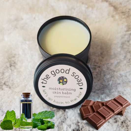 Open container of 'the good soap' moisturizing skin balm with peppermint leaves, a bottle of peppermint oil, and a chocolate bar on a textured surface.