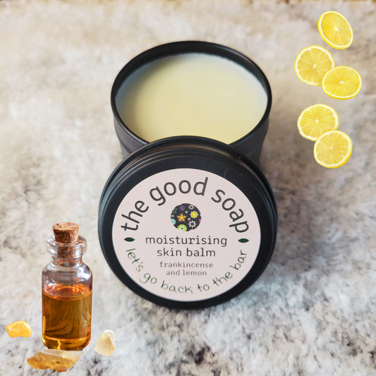 Black tin of 'the good soap' moisturising skin balm with lemon slices and frankincense oil bottle on a textured surface.