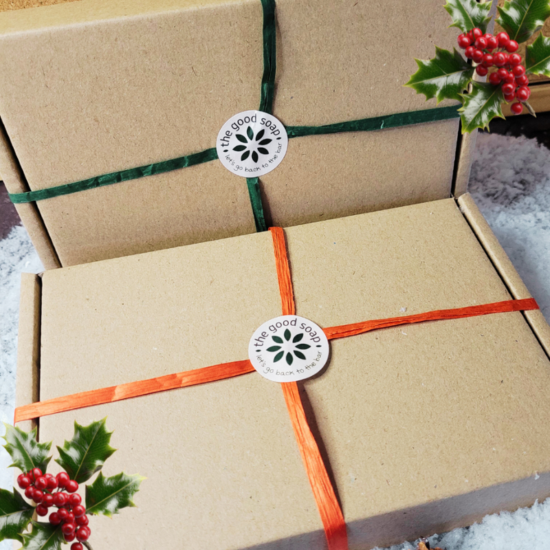 Gift box wrapped with brown paper and orange ribbon, featuring 'The Good Soap' label, surrounded by holly leaves and berries.