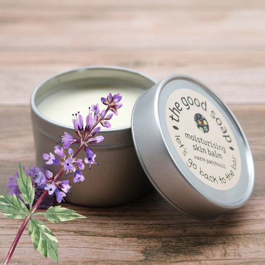 Open tin of moisturising skin balm with a label and purple flowers on a wooden surface