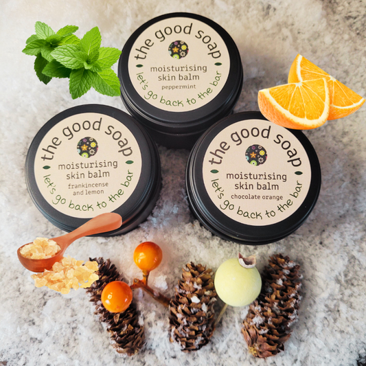Three containers of 'The Good Soap' moisturizing skin balm with natural elements on a textured surface.