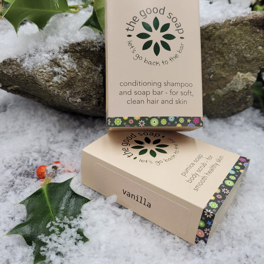 Two beige soap bars with vanilla labeling, one labeled as 'The Good Soap' and the other as 'Let's Go Back To The Bar', placed on a rocky surface with snow and greenery in the background.