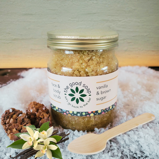 Jar of vanilla and brown sugar scrub with a wooden spoon and flowers on a textured surface.
