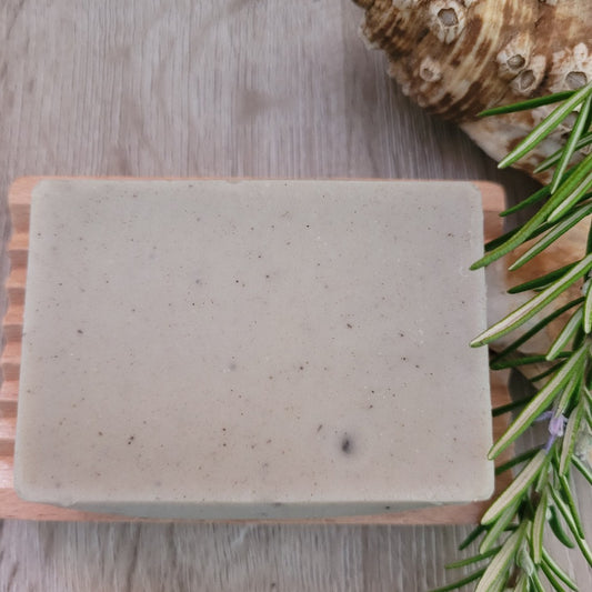 An unpackaged Mineral Mud and Rosemary Soap & Shampoo Bar  on a wooden soap dish. Bar is decorated by a sprig of rosemary