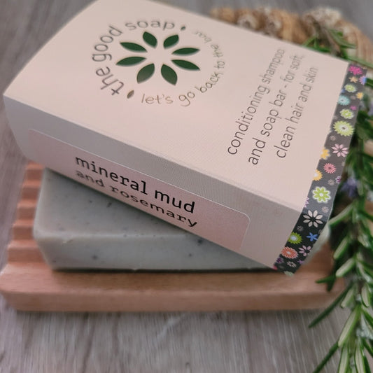 A Good Soap Mineral Mud and Rosemary Soap & Shampoo Bar in packaging on a wooden soap dish. The bar is decorated by a sprig of rosemary
