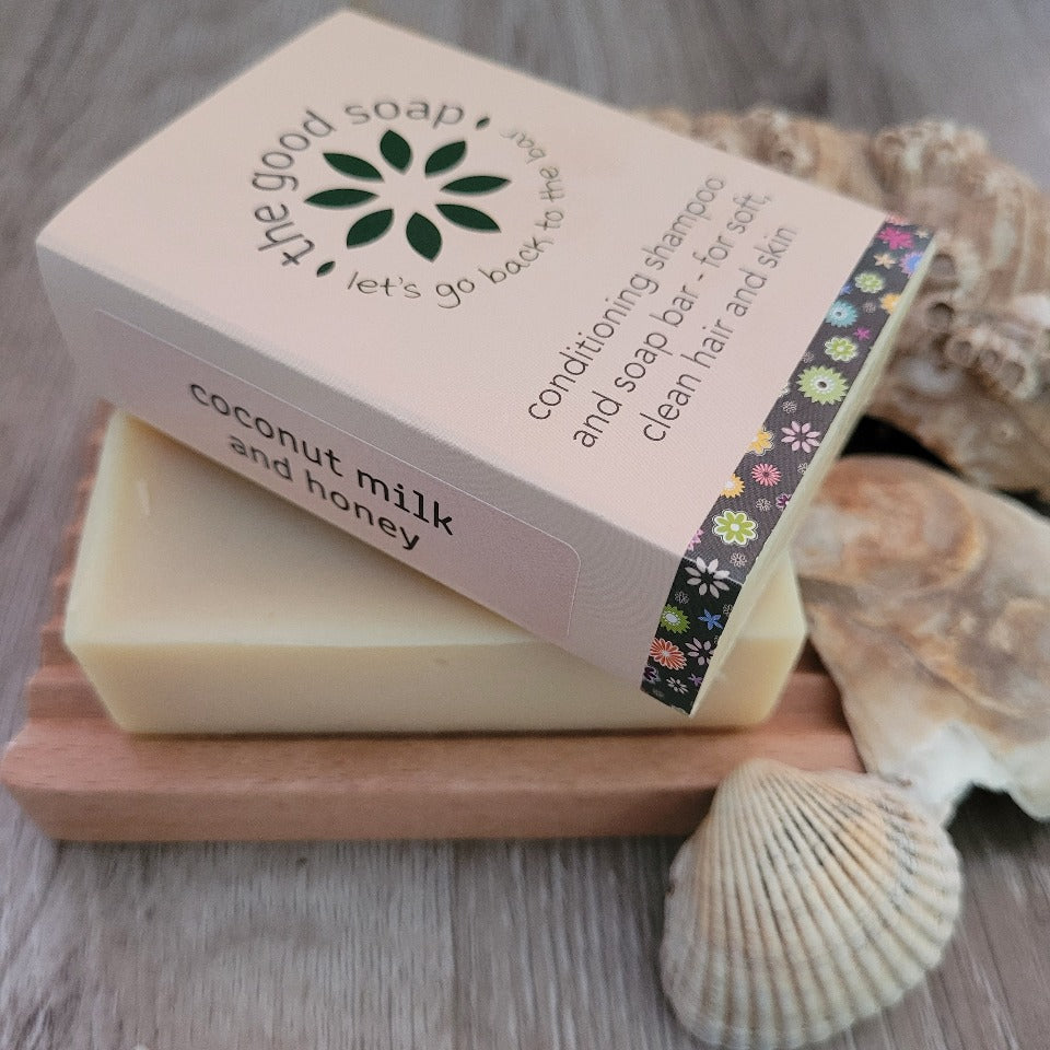 A Coconut Milk and Honey Soap & Shampoo Bar in packaging on a wooden soap dish with some shells for decoration.