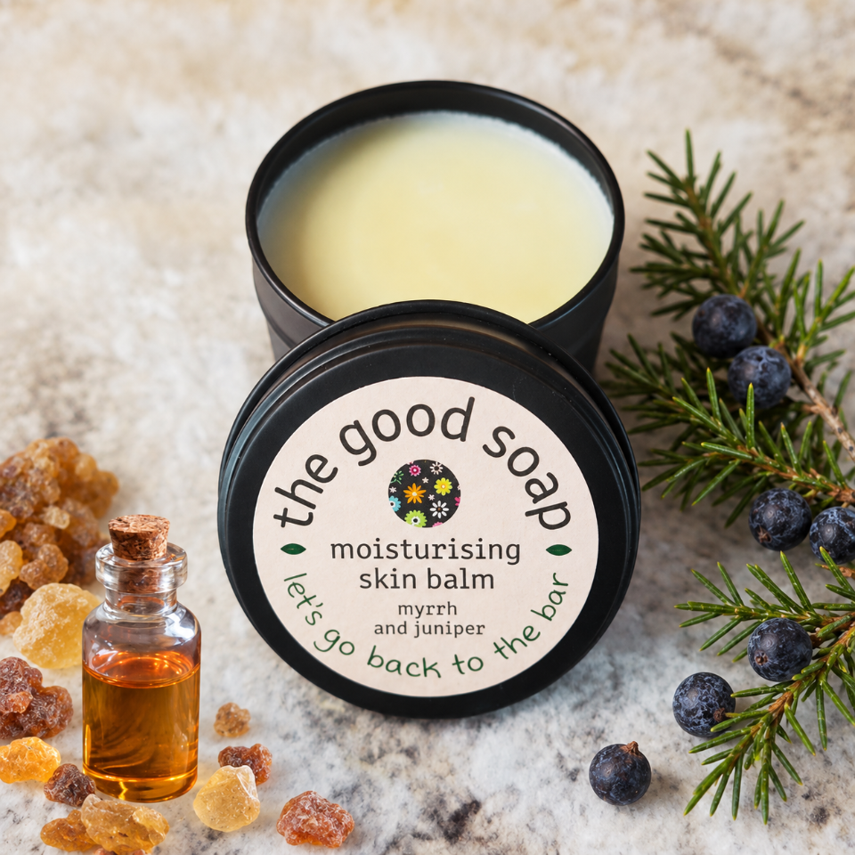 An open black tin of 'the good soap' moisturising skin balm sits on a light stone surface, with its lid placed beside it. The balm inside is pale yellow.