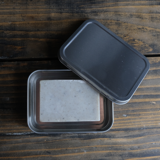 A Pumice Soap bar from The Good Soap in an open rectangle storage tin. Tin is on a wooden background.