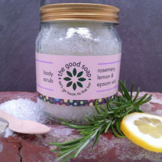 Epsom Salt Scrub with Rosemary & Lemon in a glass jar. Next to the jar is a bamboo spoon and some pieces of rosemary and lemon