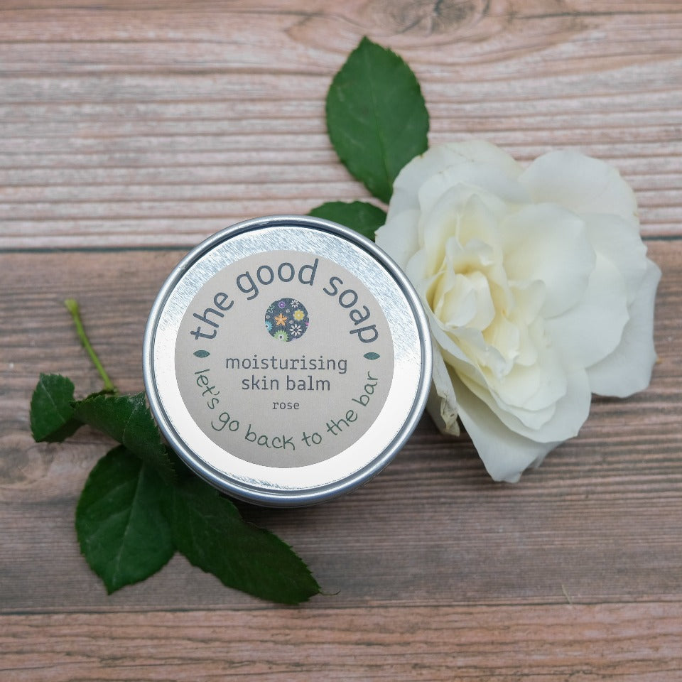 A tin of Cocoa Butter and Rose Skin Balm on a wooden background, decorated with a white rose