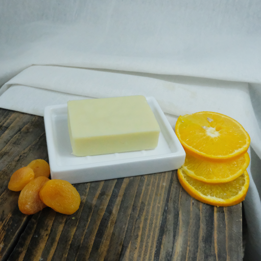 Apricot Kernel, Orange and Lavender Moisturiser Bar on a ceramic dish, with apricots and oranges for decoration. Background is a piece of muslin cloth on wood