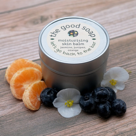 A  tin of Jasmine, Juniper and Orange Skin Balm on a wooden background. Also pictured are orange slices, pretty white flowers and juniper berries