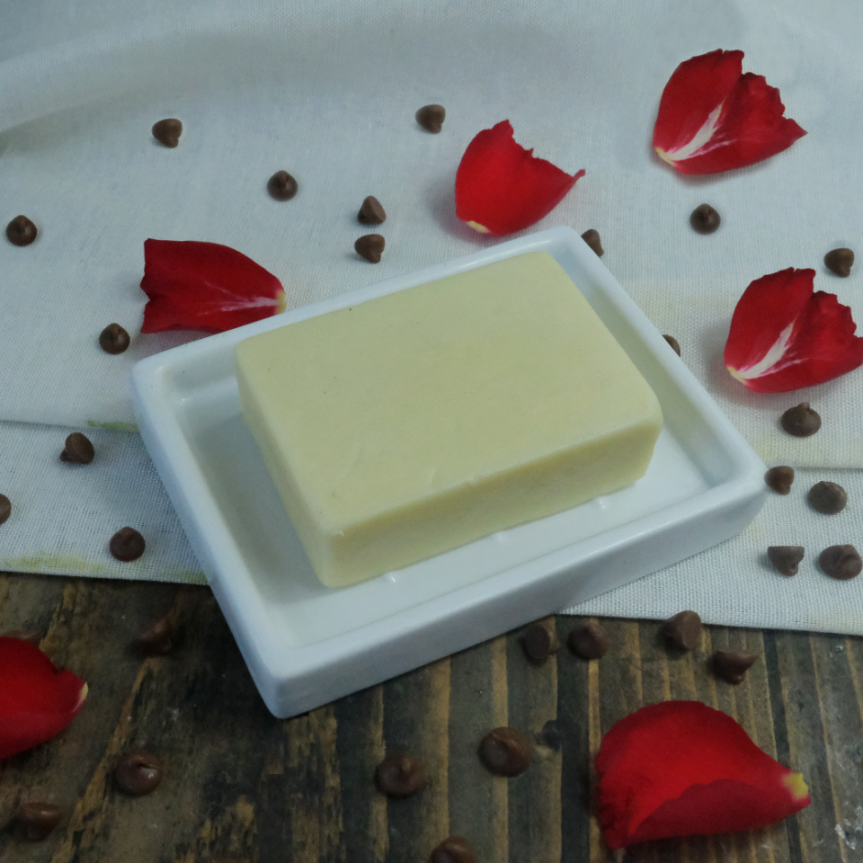 A solid moisturiser bar in Cocoa Butter & Rose scent. The bar is on a ceramic dish placed on a muslin cloth and is surrounded by rose petals and chocolate chips.