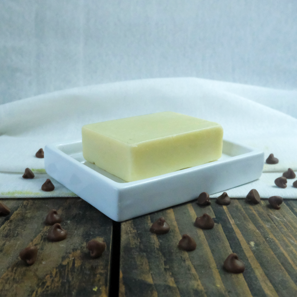 A solid moisturiser bar in Cocoa Butter & Vanilla scent. The bar is on a ceramic dish on a wooden background and is surrounded by chocolate chips.