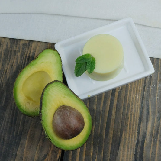 Avocado Butter and Mint Moisturiser Bar Foot Balm on a ceramic dish, decorated with a piece of fresh mint and a halved avocado. Bar is on a wooden background on a piece of muslin cloth